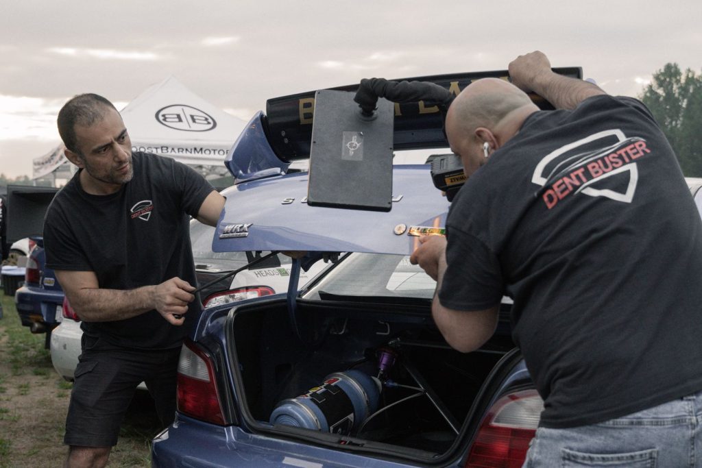 Two technicians reading dents on a blue Subaru trunk lid under a light board