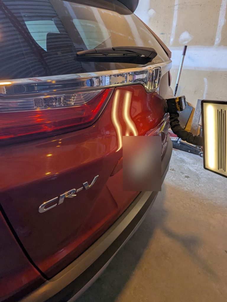 Dent above the plate on a red Honda CR-V tailgate, reflection lines bent under the PDR light
